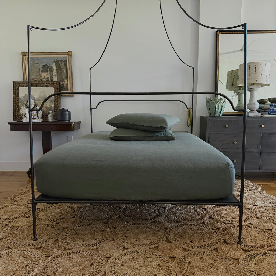 A black metal four-poster bed frame with a minimalist arched headboard and footboard, styled with sage green bedding and pillows on a woven jute rug in a bedroom setting.